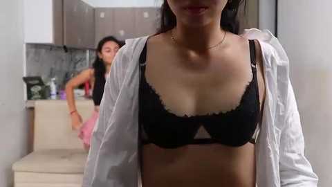 Video of an Asian woman in a black bra, white shirt, standing in a modern kitchen with beige cabinets. Another woman, partially visible, is cooking in the background.