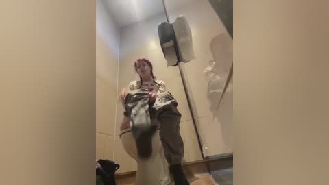 Video of a person in a public restroom, sitting on a toilet, wearing a white t-shirt with red and black patterns, and gray pants. The restroom has beige walls and a mirror, with a paper towel dispenser above the toilet.