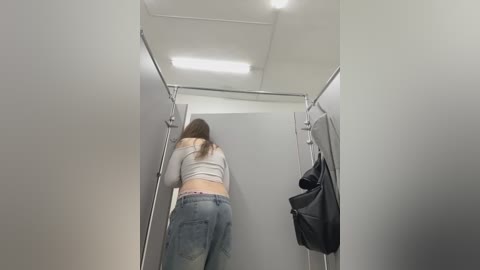 Video of a woman in a white crop top and blue jeans walking away from the camera in a minimalist, bright, white-walled dressing room with a black handbag hanging on a hook.
