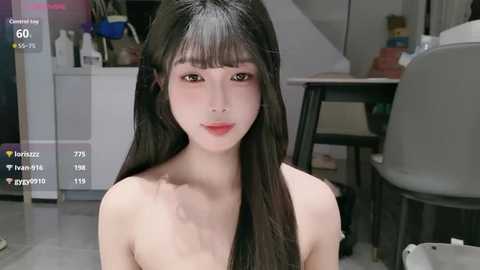 Video of an East Asian woman with long black hair and fair skin, wearing minimal makeup, sitting in a modern, sparsely furnished room with a white countertop and chairs.