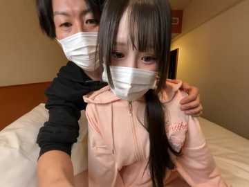 Video of two Asian women in face masks, one hugging the other, both in pink hoodies, in a dimly lit room with beige walls and a bed.