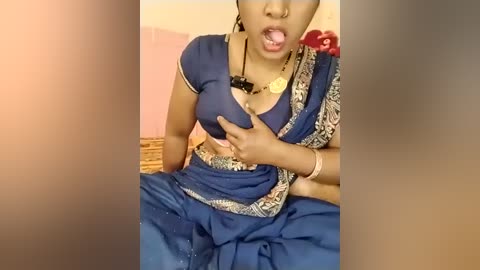 Video of a South Asian woman wearing a blue sari with intricate designs, pressing a breast with her left hand, tongue out, and a necklace visible. Background shows a pink wall and a red flower.