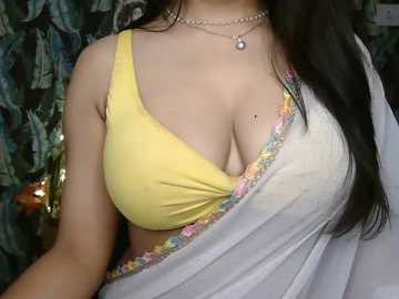 Video of a woman with light brown skin, wearing a yellow bra, a white sari with colorful floral border, and multiple necklaces, against a blurred green and blue floral background.