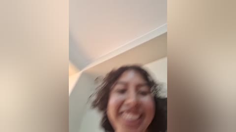 A blurry video of a woman with dark hair, smiling broadly, taken from a low angle, partially obstructed by a white wall. The background is out of focus.