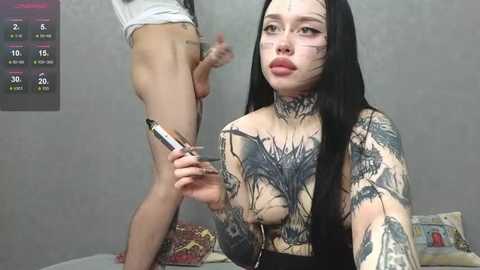Video of a tattooed woman with long black hair, holding a camera, in a bedroom with a gray wall and a bed with colorful pillows. A naked man stands beside her.