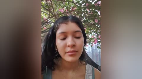 Video of a young woman with curly black hair and medium skin tone, wearing a striped tank top, standing under a tree with pink flowers.
