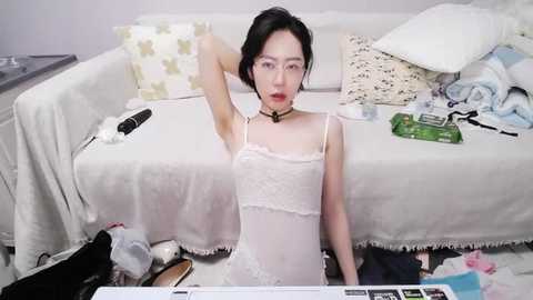 Video of an East Asian woman with short black hair, wearing a sheer white lace nightgown, sitting on a messy bed with scattered clothes and electronics, in a cluttered bedroom.