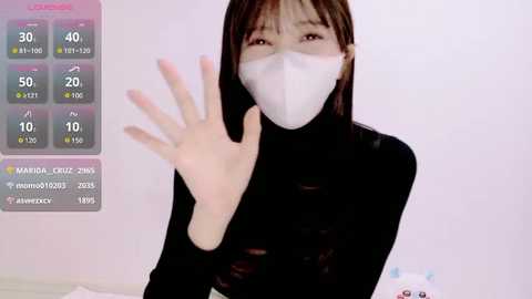 A video of an East Asian woman with straight, shoulder-length brown hair, wearing a white face mask and black long-sleeve top, waving her hand in a video call. Background shows a white wall with a virtual weather display.