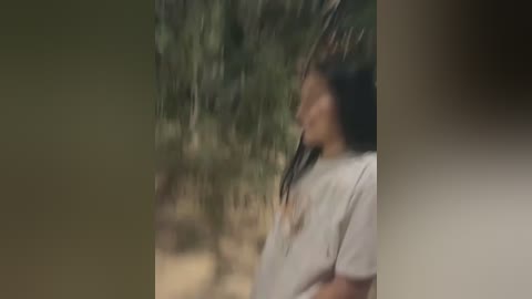 A blurred video of a person with long, dark hair, wearing a white t-shirt, standing amidst greenery. The background is out of focus, suggesting a natural outdoor setting. The image is soft and slightly grainy.
