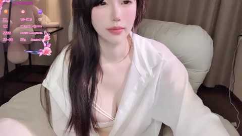 Video of a pale-skinned woman with long, dark hair, wearing a sheer white robe, revealing a beige bra, sitting on a beige couch in a dimly-lit room with floral decorations.