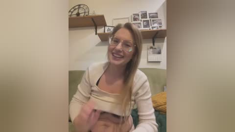 Video of a smiling, fair-skinned woman with long blonde hair, wearing glasses and a beige cardigan, lifting her shirt to reveal her midriff and small breasts. She sits on a green couch in a cozy, beige-walled room with shelves and framed photos.