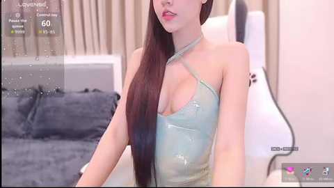 A video of an East Asian woman with long, straight brown hair, wearing a translucent, light blue halter top, seated in a modern bedroom with beige curtains and a gray bed.