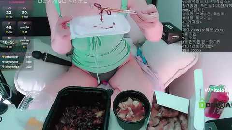 Video of a person in green lingerie, sitting cross-legged, holding a white box, with a black container of food and a pink bottle on a table.