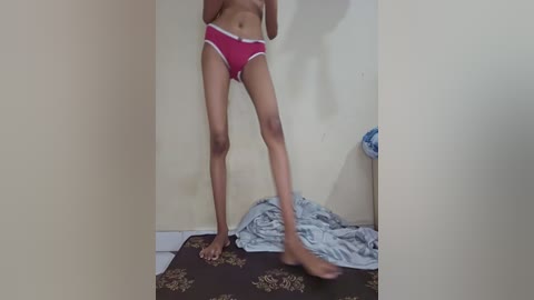 Media: Video of a slender, light-skinned person wearing red panties with white trim, standing in a cluttered room with a beige wall and a carpeted floor.