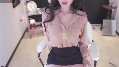 Video of an Asian woman with long black hair, wearing a beige blouse with a zipper, black shorts, and a green name tag, seated in a white chair in a modern room with white walls and dark furniture.