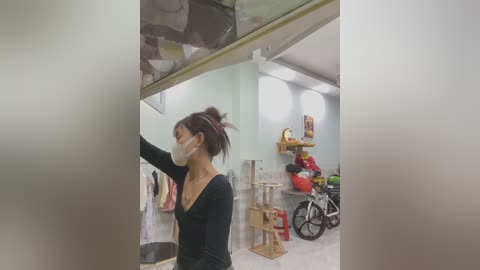 Video of an Asian woman with dark hair in a ponytail, wearing a black long-sleeved top, face mask, and surgical gloves, inside a sterile room with a ceiling fan, a white bicycle, and various medical supplies.