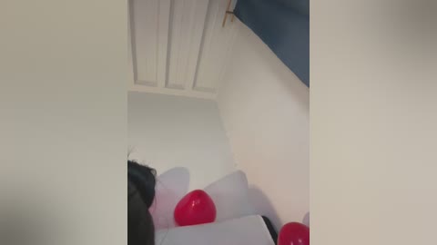 Video of a narrow, dimly lit room with white walls, a sloped ceiling, and three red balloons. A black jacket is partially visible. The room has a minimalist, clutter-free appearance.