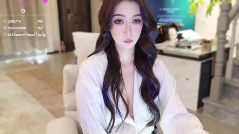 A video of an Asian woman with long, wavy black hair, wearing a white button-down shirt, seated in a modern living room with a large TV and plants.