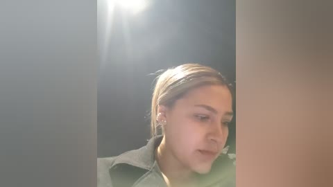 Video of a young woman with light skin, blonde hair, and a neutral expression, wearing a dark jacket, standing in a dimly lit room with a bright light source above.