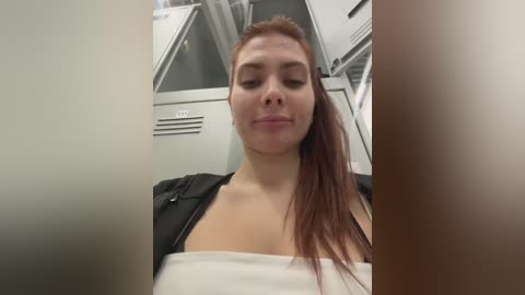 Video of a young Caucasian woman with light skin and long red hair, wearing a black jacket over a white top, inside a dimly lit airplane cabin with overhead storage bins in the background.