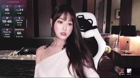Video of an East Asian woman with long black hair, wearing a white off-shoulder top, standing in a dimly lit room with a gaming chair and desk in the background.
