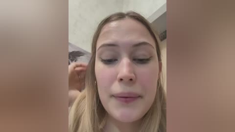 Video of a young woman with long blonde hair, light skin, and closed eyes, being combed by an unseen person. The background shows a beige wall and part of a white ceiling.