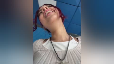 A video of a smiling woman with short, vibrant red hair, wearing a white top and black necklace, against a blue and white ceiling background.