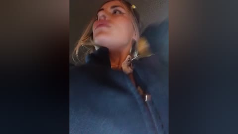 Media: Video of a young woman with blonde hair, wearing a black coat, looking upward with a neutral expression. The background is blurred, emphasizing her face.
