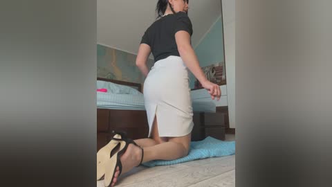 Video of a woman with light skin, wearing a black top and white pencil skirt, kneeling on a bed with a blue blanket. She has black high-heeled shoes. The room has a map on the wall and a wooden dresser.