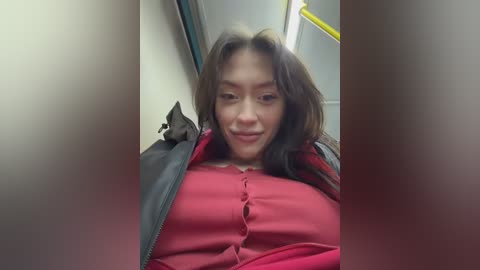 A video of an Asian woman with shoulder-length dark hair, wearing a red blouse and black jacket, sitting on a subway bench, smiling, with blurred background.