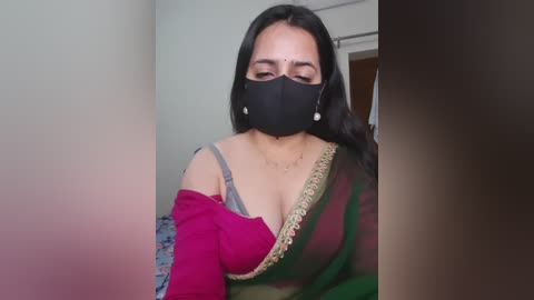 Media: Video of a South Asian woman with medium skin tone, long black hair, wearing a pink off-shoulder blouse, green sari, black face mask, and dangling earrings. Background features a neutral-colored room with a door.