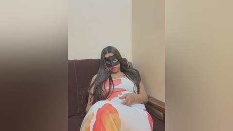 Video of a South Asian woman with long dark hair, wearing a white dress with red and orange floral patterns, black mask, and seated on a brown couch in a dimly lit room with beige walls.