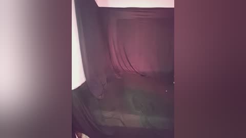 A video of a dimly lit room with a draped window and a faintly visible figure sitting in the foreground, partially obscured by shadows and soft lighting.