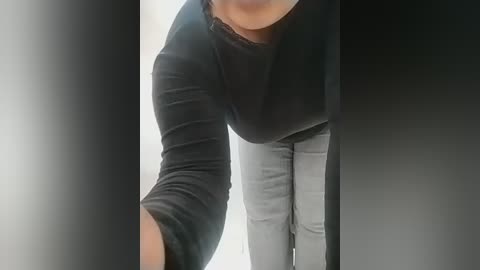 A close-up video of a person with a medium skin tone, wearing a black long-sleeved shirt and gray pants, bending forward, possibly taking a selfie.