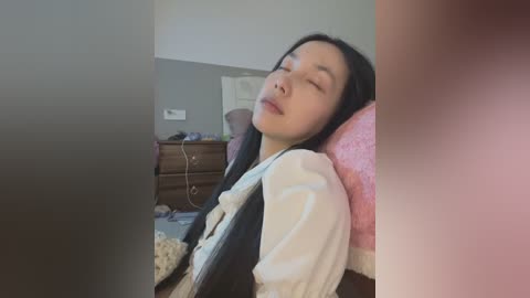Media: A video of an East Asian woman with long black hair, closed eyes, and a relaxed expression, wearing a white blouse and black vest, sitting in a pink cushioned chair in a dimly lit, cluttered room.
