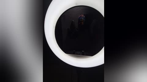 A video of a dark, dimly lit room with a circular mirror and bright white light from the ring light, creating a halo effect. A colorful, patterned hat is visible inside the mirror.