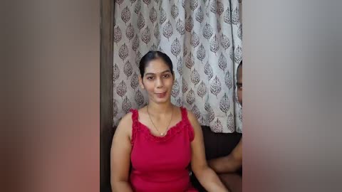 Video of a woman with medium brown skin and dark hair tied back, wearing a red sleeveless top, seated in a room with patterned curtains.