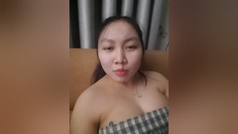 Video of an Asian woman with fair skin, long dark hair, and light makeup, sitting topless on a brown couch. She has visible sweat and a checkered blanket covers her chest. Background includes silver curtains and a blurred wall.