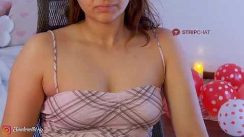 Video of a light-skinned woman with medium brown hair, wearing a pink plaid camisole, sitting indoors. Background includes red and white polka-dotted balloons and a lit candle.