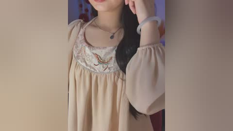 Video of a young woman with long black hair, wearing a beige blouse with floral embroidery, a silver necklace, and a white scrunchie, against a blurred background.