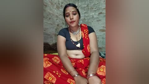Video of an Indian woman with medium skin tone, dark hair, wearing a red sari with yellow patterns, a black crop top, and gold jewelry, seated in a rustic room with a stone wall background.