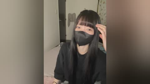 A video of an Asian woman with long black hair and bangs, wearing a black face mask, black top, and sitting on a bed with beige sheets. The room has a plain, minimalist interior with a closed door and a patterned wall.