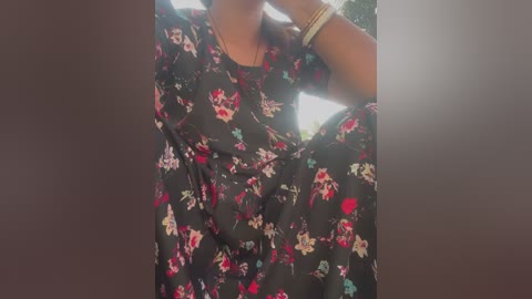 Media: Video of a woman with medium-dark skin tone, wearing a black floral dress, adjusting her hair with both hands, in a natural outdoor setting with sunlight filtering through.