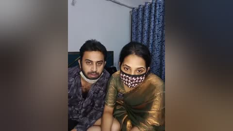 Media: Video of two South Asian men in traditional attire with masks; one in a dark patterned shirt, the other in a gold sari, seated in a room with blue curtains and a plain white wall.