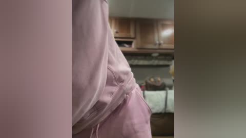 Media: Video of a person wearing a light pink apron, standing in a dimly lit kitchen with wooden cabinets, a countertop, and a partially visible stove in the background.