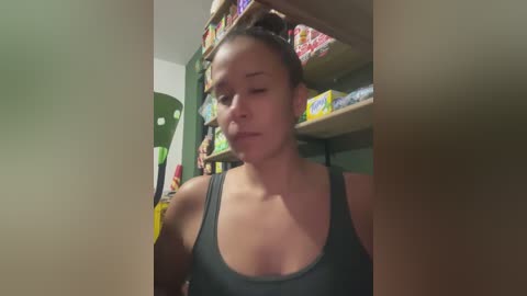 Video of a young woman with light brown skin, brown hair tied back, wearing a black tank top, standing in a brightly lit grocery store aisle with shelves filled with various products.