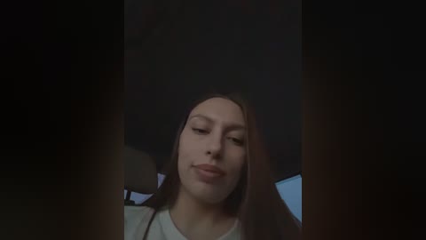 Video of a young woman with long brown hair and fair skin, wearing a white shirt, seen from the neck up, with dark, blurred background.