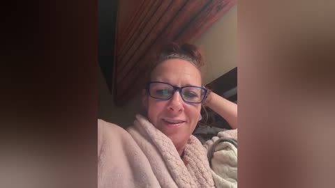 Video of a middle-aged woman with glasses, fair skin, and brown hair, wrapped in a beige towel, smiling in a cozy, dimly-lit room with wooden beams and a textured ceiling.