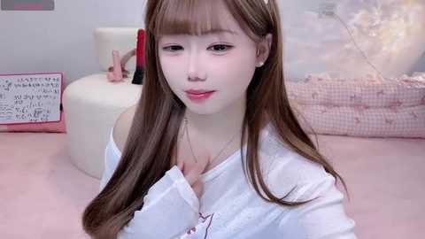 Video of a young East Asian woman with long brown hair and fair skin, wearing a white blouse, sitting on a pink bed in a softly lit room with pastel decor.