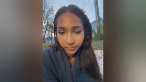 Video of a young Black woman with medium-brown skin, long dark hair, and a nose ring, wearing a dark jacket, standing outdoors with suburban houses and trees in the background.